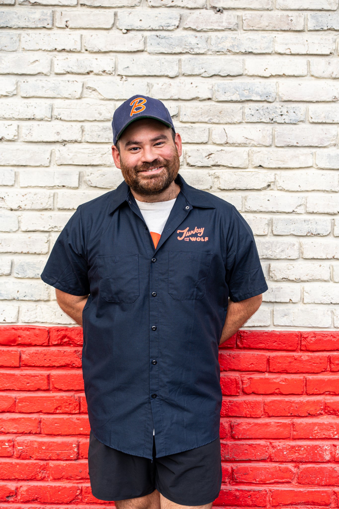 Man wearing a navy blue shirt with a logo and cap, standing against a white brick wall with red base.