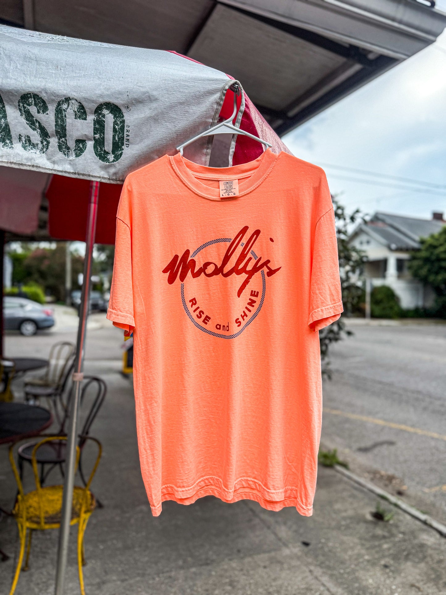 Orange t-shirt with a logo hanging on a street corner with a building and car in the background.