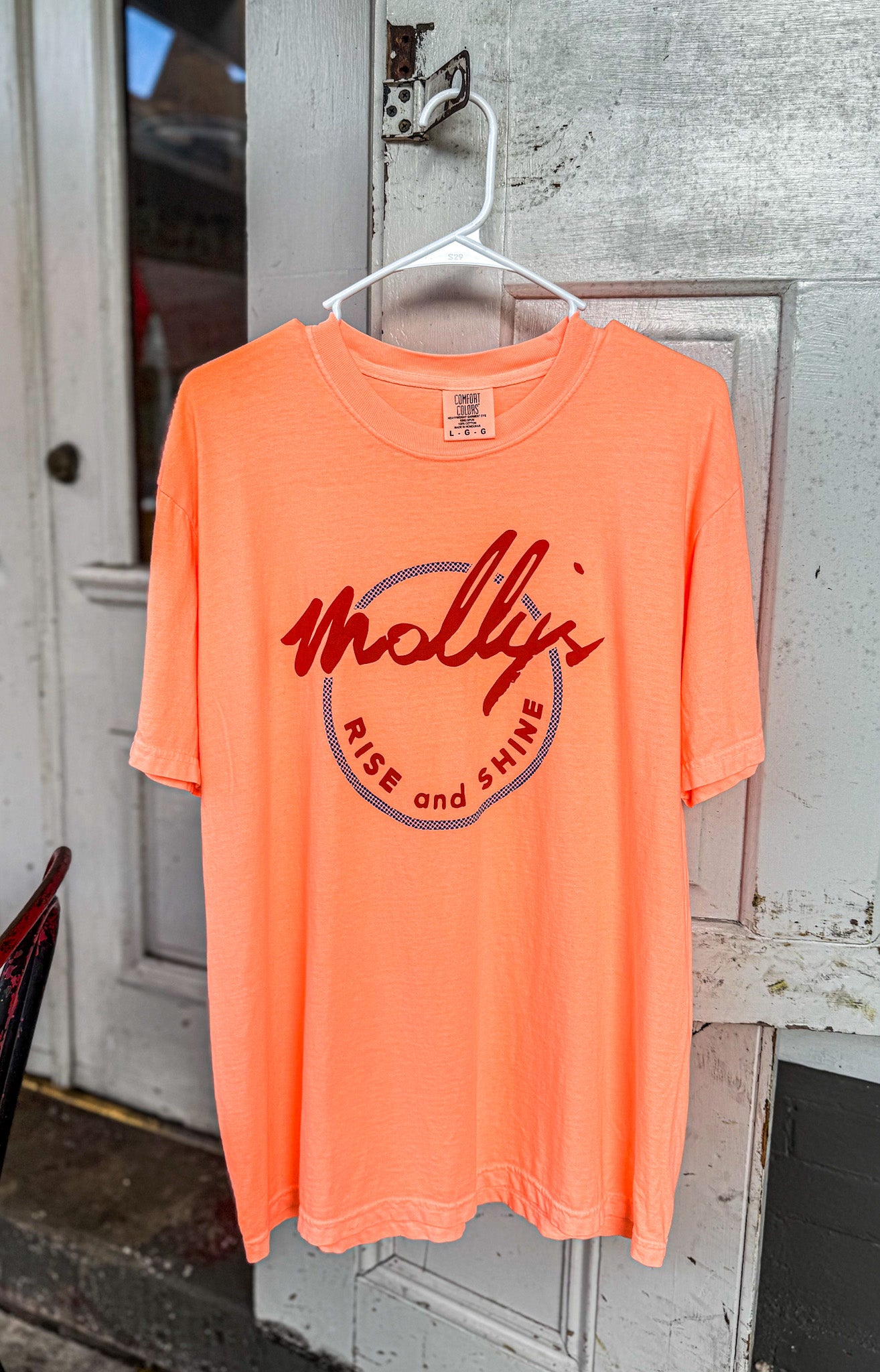 Orange t-shirt with 'Molly Rise and Shine' text on a hanger against a wooden door background