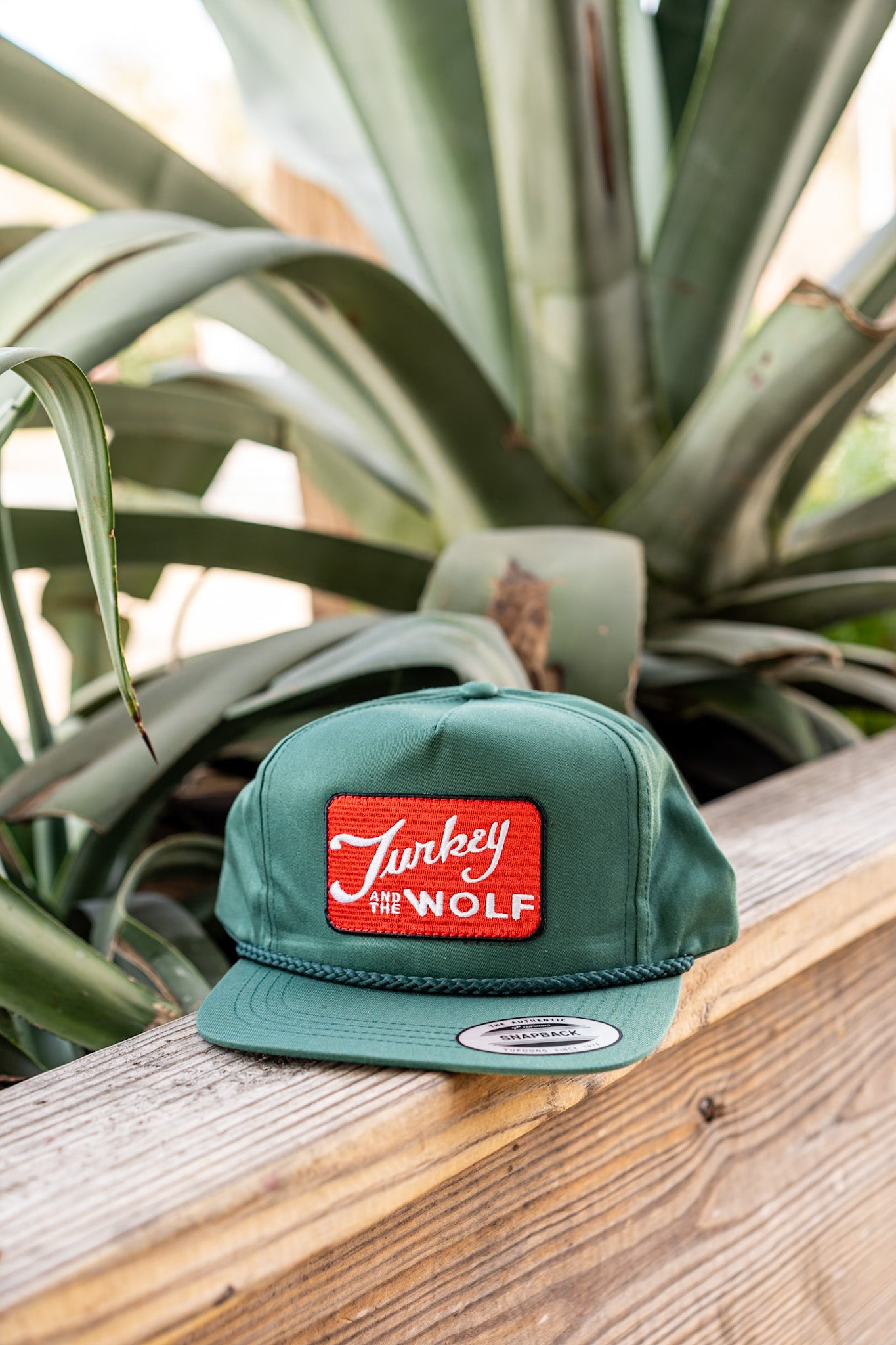 Green cap with a red patch featuring 'Turkey the Wolf' on a wooden surface with plants in the background
