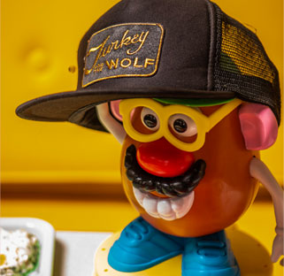 A toy figure wearing a Turkey and the Wolf cap, set against a yellow kitchen background.