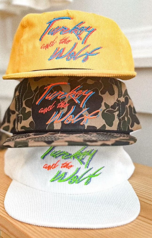 Photo of 3 hats that all say turkey and the wolf in blue and red. The hats from bottom to top are white corduroy, camo, and yellow corduroy.