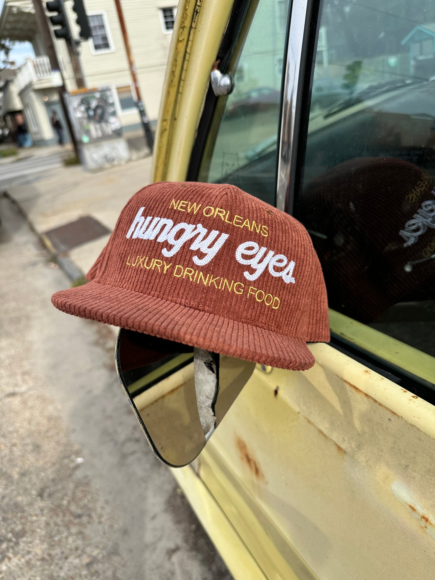 Brown corduroy hat that says hungry eyes in bold white letters, above hungry eyes it says new orleans in gold and below hungry eyes it says luxury drinking food.