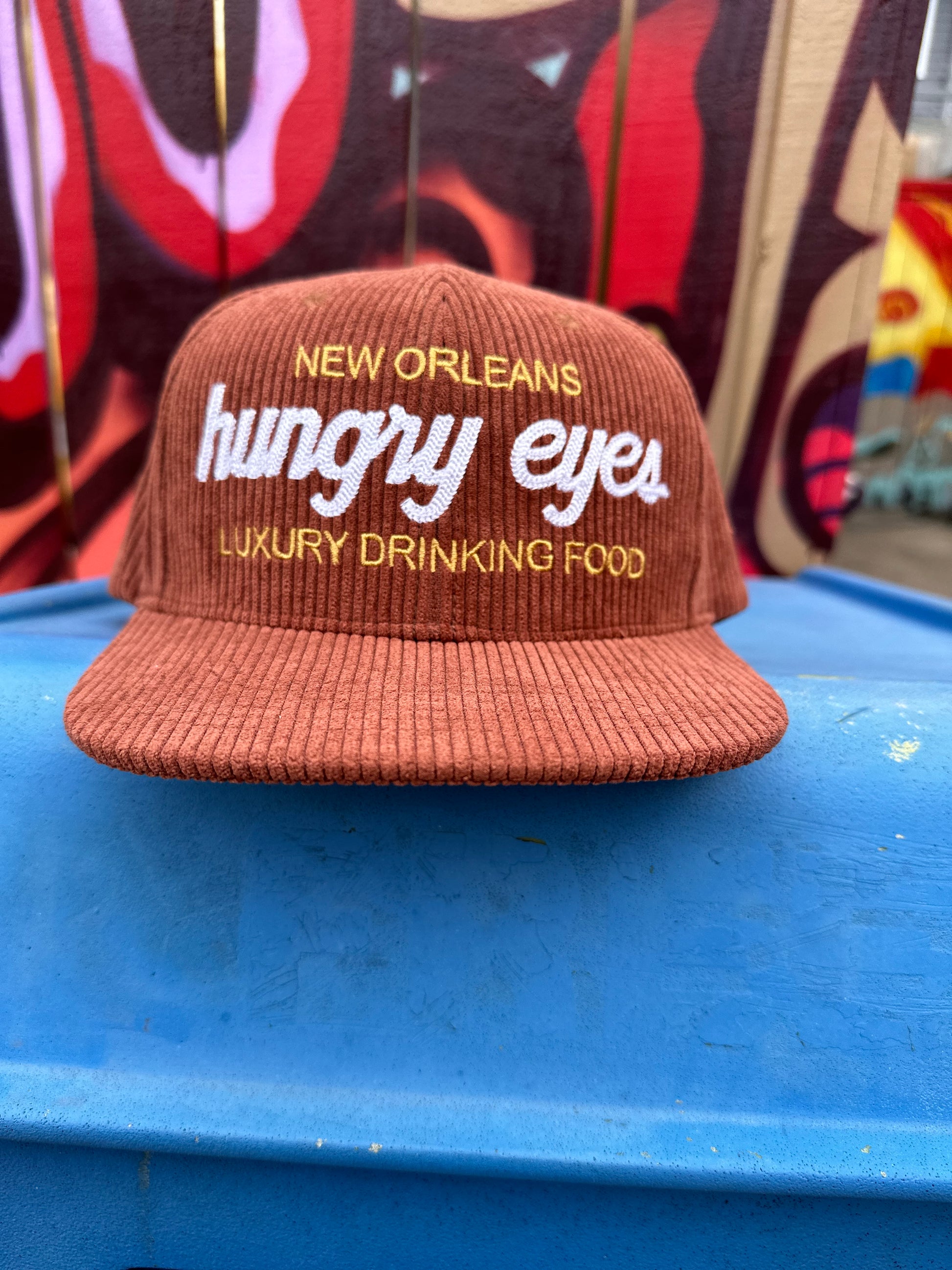 Brown corduroy hat that says hungry eyes in bold white letters, above hungry eyes it says new orleans in gold and below hungry eyes it says luxury drinking food.