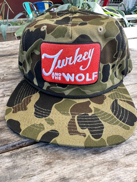 A camouflage-print Turkey and the Wolf cap on a wooden table, outdoors in daylight.