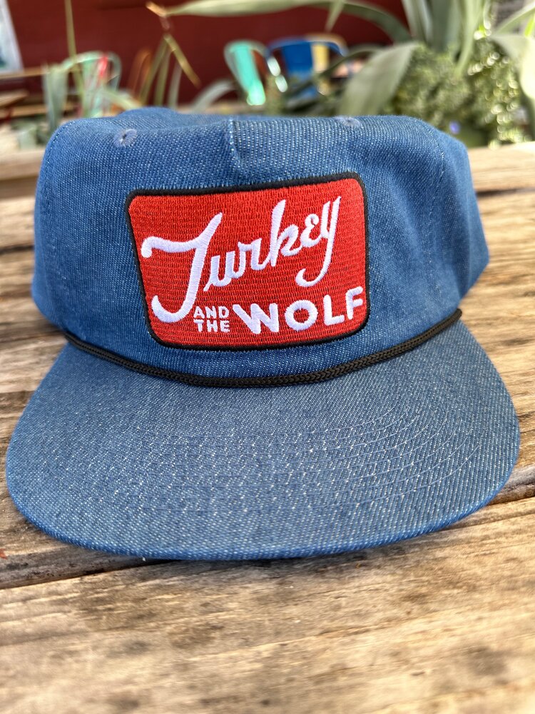 A denim blue Turkey and the Wolf cap with a red patch on a wooden table in an outdoor setting.
