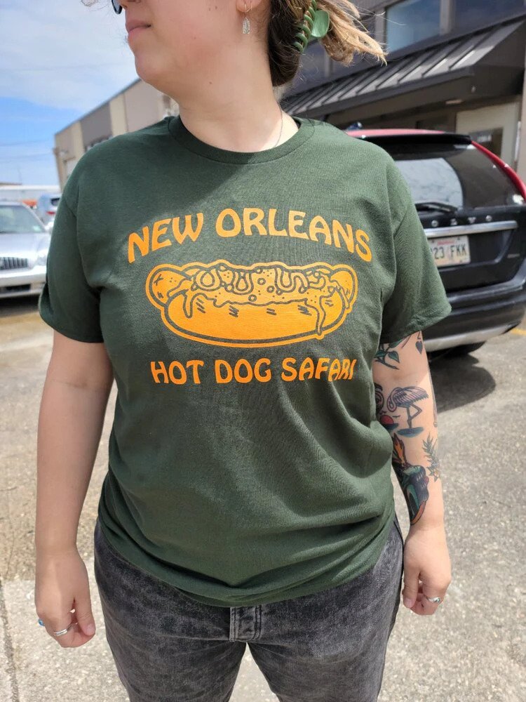 Person in a green "New Orleans Hot Dog Safari" t-shirt, with a partial view of their face and tattooed arm