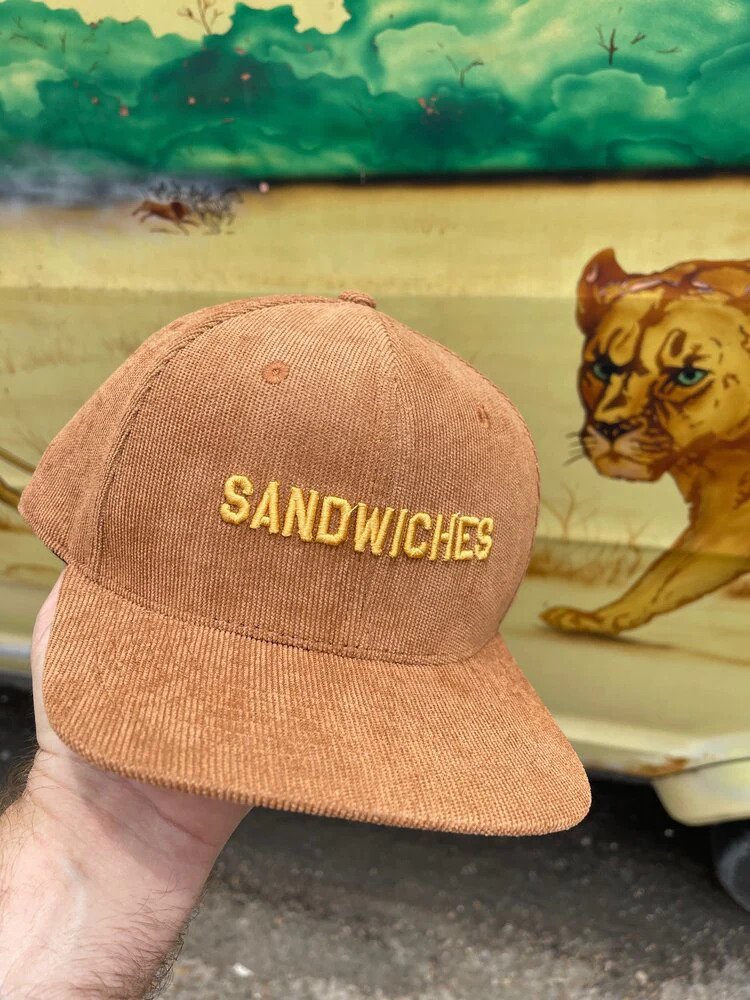 A corduroy cap with the word SANDWICHES in front of a mural featuring a lion.