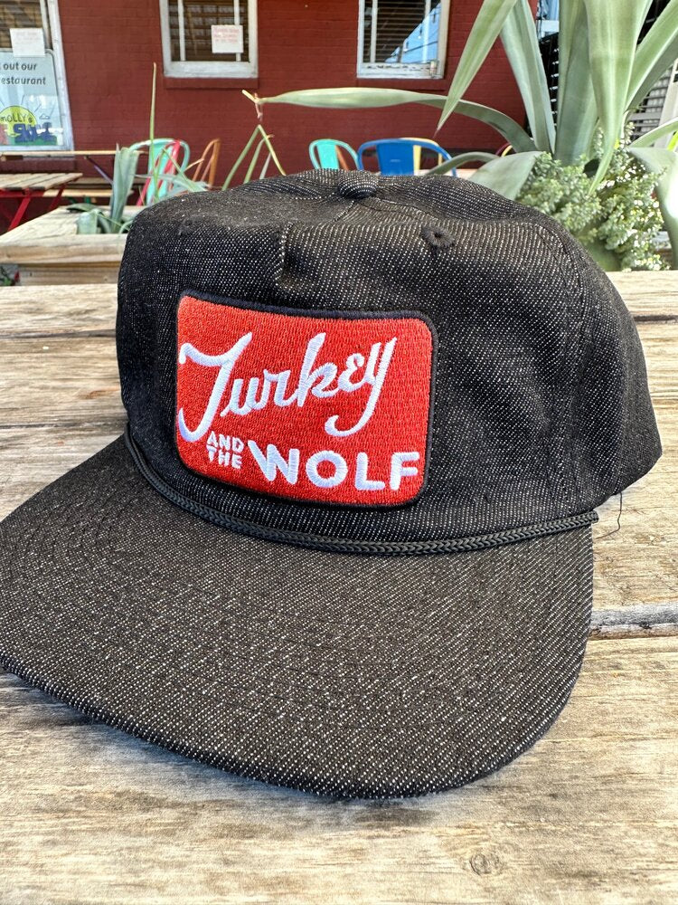 A black textured Turkey and the Wolf cap with a red logo on a rustic wooden surface outdoors.