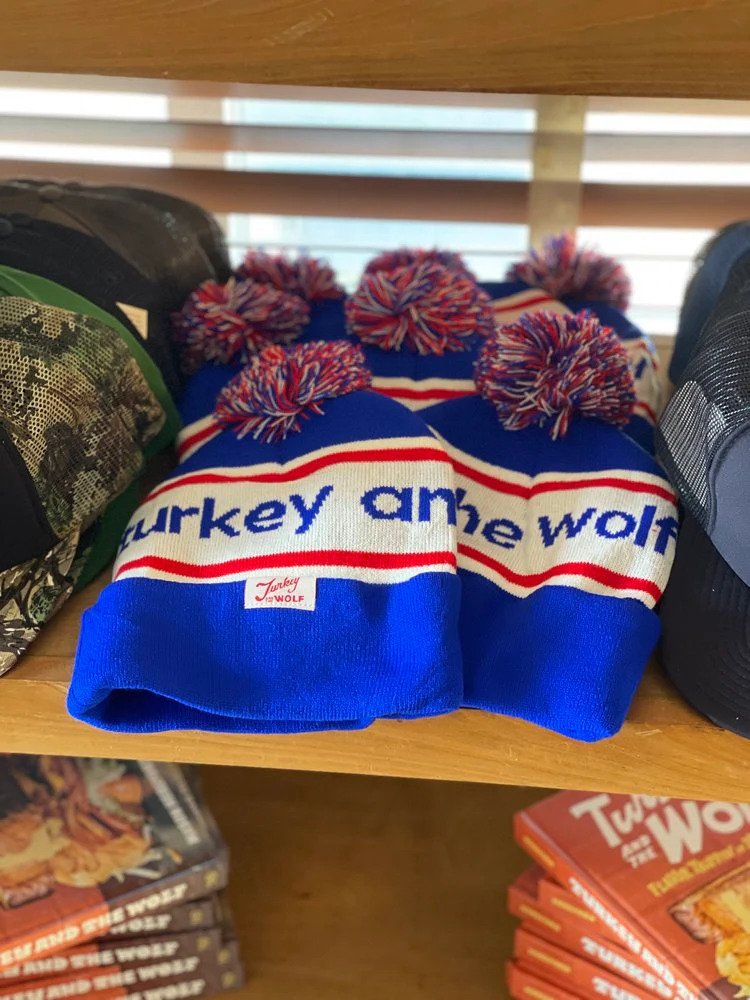 Blue and white striped knit hats with pom-poms and the text Turkey and the Wolf displayed on a shelf, with books below.