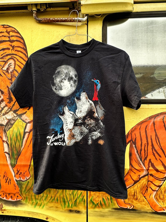 Black T-shirt with  2 wolves and 1 turkey looking at a full moon with a starry sky background. Shirt says turkey and the wolf on the lower left corner of the design. T-Shirt is on a hanger in front of a vehicle with tigers on it.