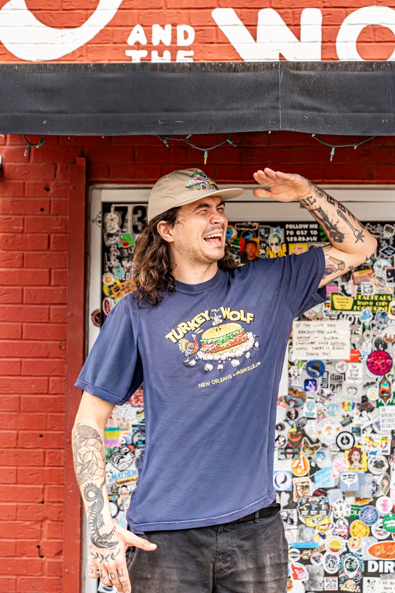 Man wearing a blue t-shirt with a graphic design, standing in front of a brick wall with stickers.