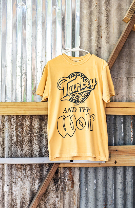 Yellow t-shirt with 'Turkey and the Wolf' design on a wooden surface with metal background