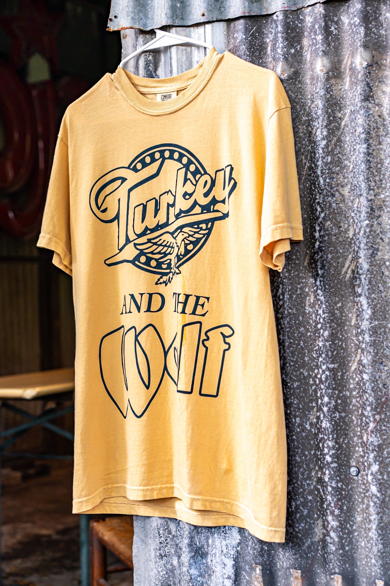 Yellow t-shirt with graphic design hanging against a corrugated metal wall.