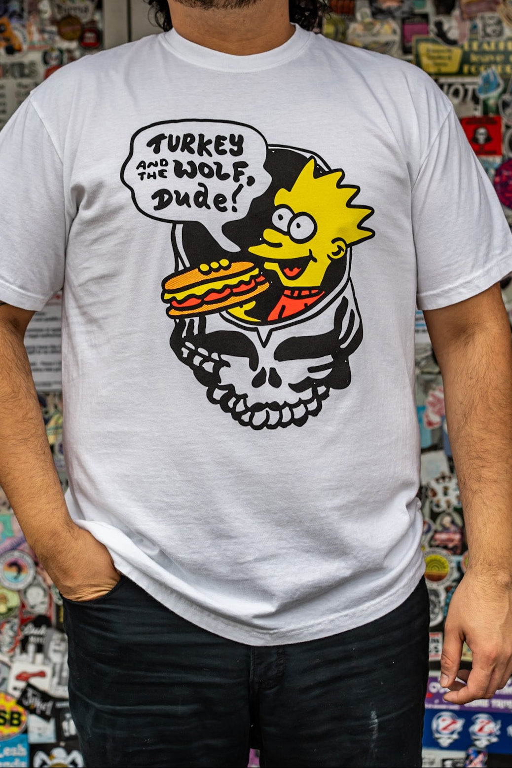 Person wearing a white t-shirt with a graphic design in front of a wall covered in stickers.