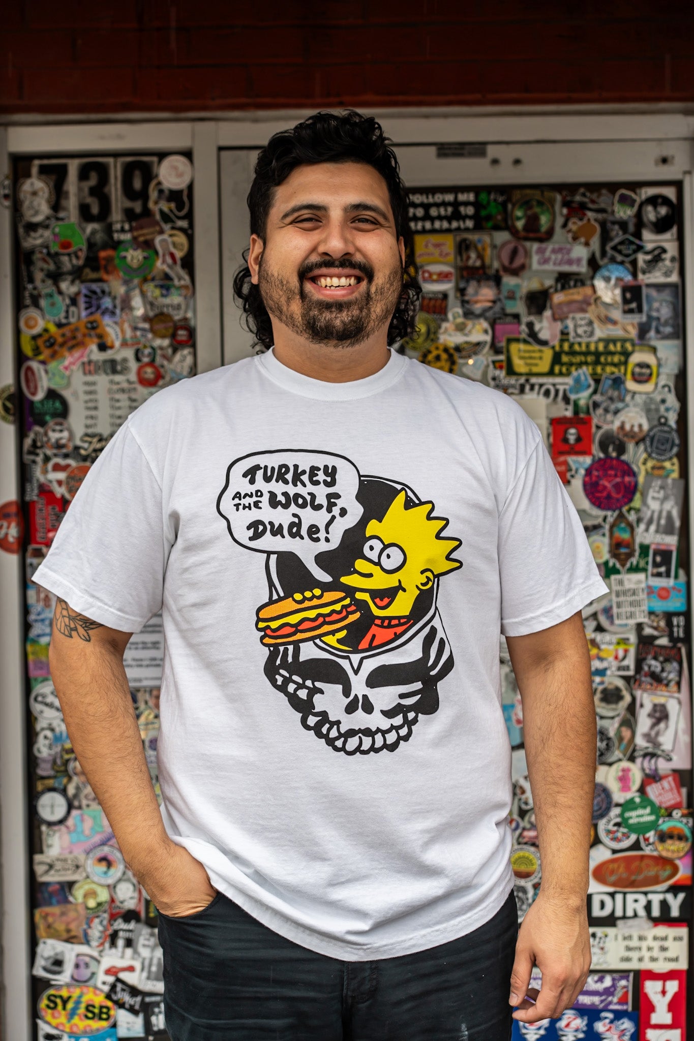 Man wearing a white t-shirt with a graphic design in front of a wall covered in stickers.
