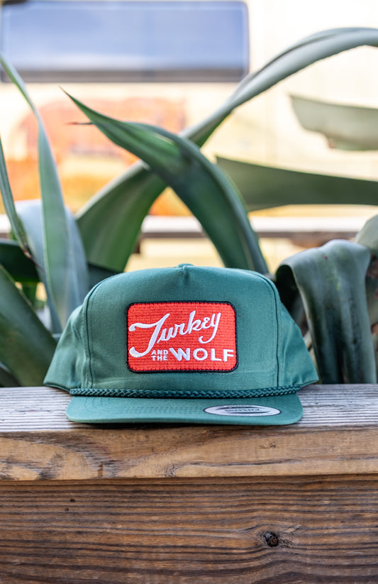 Green cap with a red patch featuring 'Turkey the Wolf' on a wooden surface with plants in the background