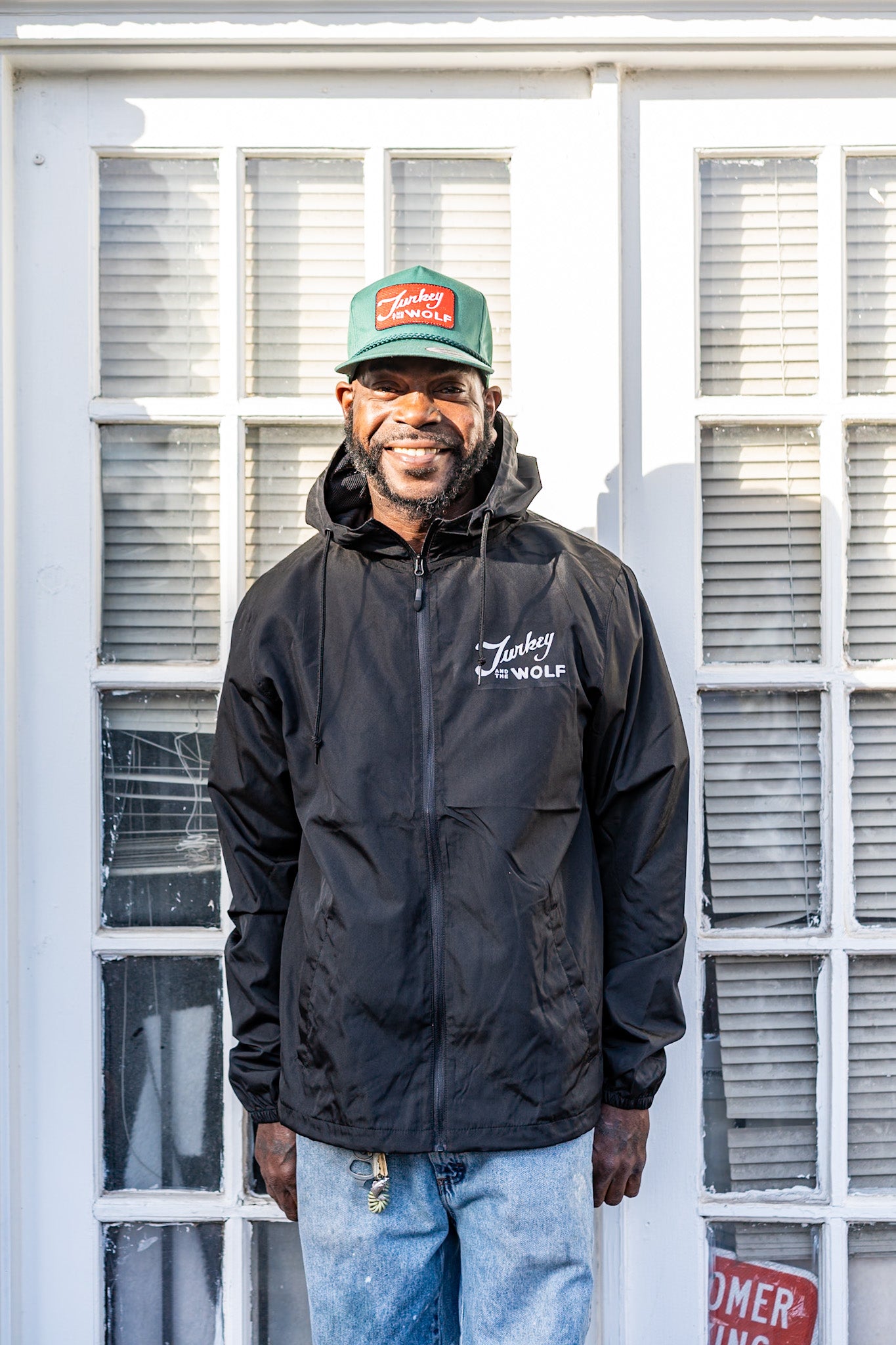Man wearing a black jacket and green cap standing in front of a white door with glass panels.