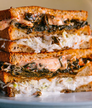Stacked sandwich on a plate with creamy coleslaw and dark leafy greens, toasted bread layers visible.