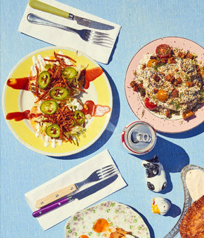 Sunlit dining table with plates of salad, cutlery set, a can of soda, and a sandwich, on a blue cloth background.
