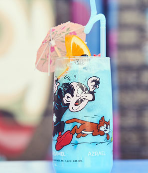 Tall glass with a blue drink and garnished with orange and umbrella, featuring a cartoon character, against a blurred background.