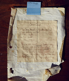 An aged, handwritten recipe for corn chowder taped to a wooden surface, with a blue sticky note.