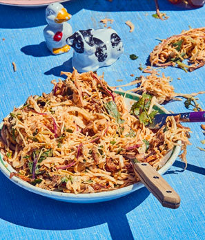 A bowl of shredded salad with utensils on a bright blue table, in sunny outdoor setting.