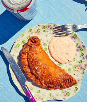 A fried fish fillet with sauce on a decorative plate, with cutlery and a soda can, on a blue surface.