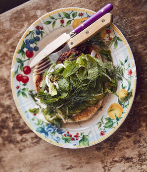 Herb-topped dish on a floral plate with fork and knife on a rustic wooden table.
