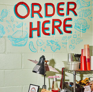 A restaurant counter with an ORDER HERE sign, doodles on the wall, and various items on the counter.