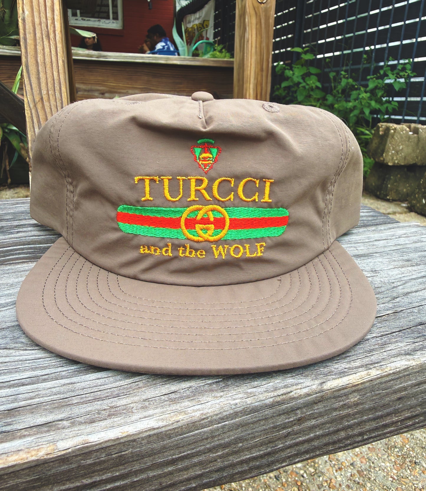 A brown flat-brim baseball cap with embroidered 'TURCCI and the WOLF' text and a green and red stripe design on the front, displayed on a weathered wooden surface outdoors.