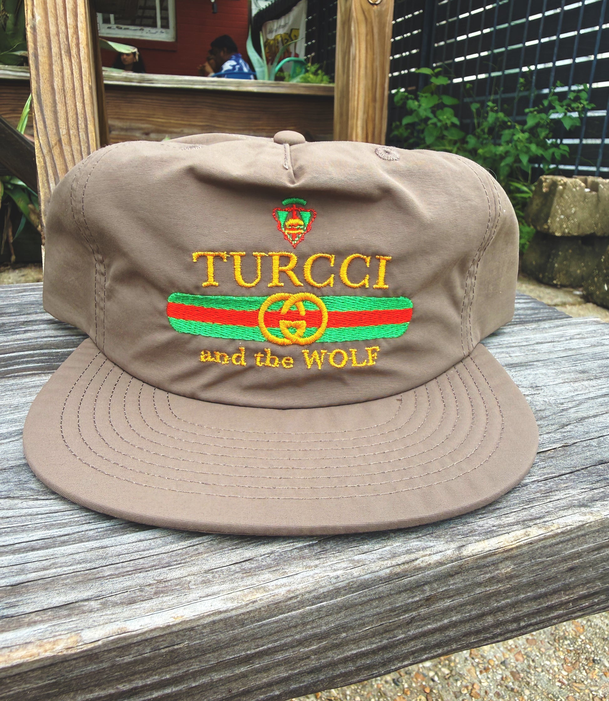 A brown flat-brim baseball cap with embroidered 'TURCCI and the WOLF' text and a green and red stripe design on the front, displayed on a weathered wooden surface outdoors.
