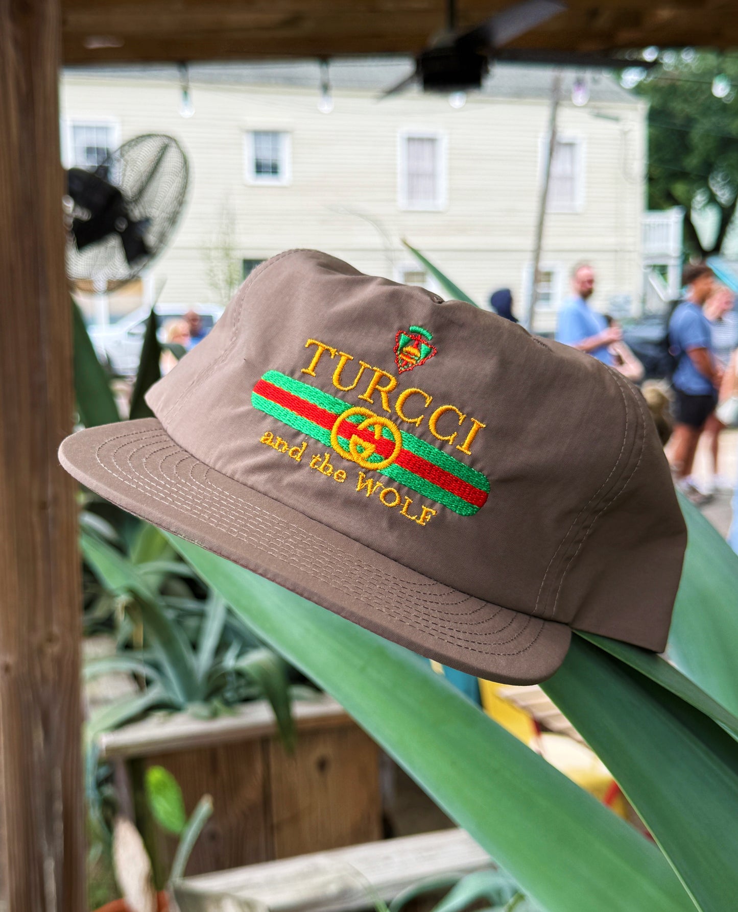 A brown flat-brim cap with embroidered 'TURCCI and the WOLF' text and a red-green-red stripe design, displayed outdoors on a green leaf with a blurred background of people and a building.