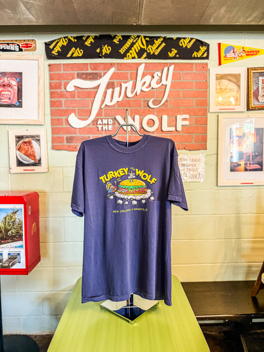 Blue t-shirt with 'Turkey and the Wolf' graphic on a mannequin against a wall with framed pictures and a sign.