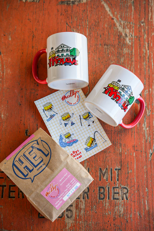 Two mugs with colorful designs on a wooden surface, accompanied by a small bag labeled 'Hey'.
