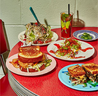 A retro diner table laden with assorted sandwiches and salads, complete with a whimsical glass and straw, against a red backdrop.