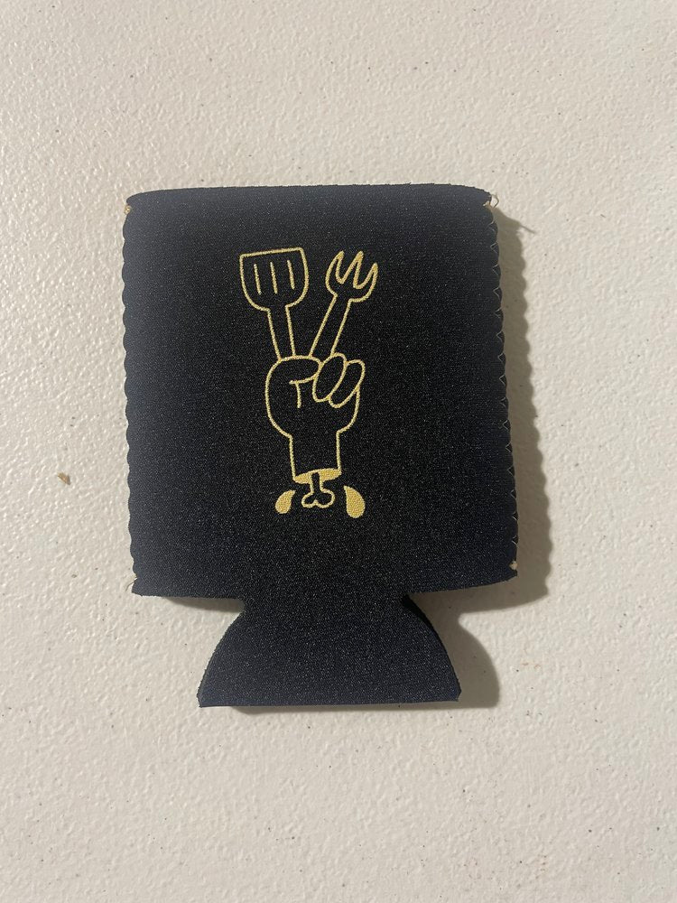 A black beverage insulator with a gold utensil-holding hand graphic, against a white wall.