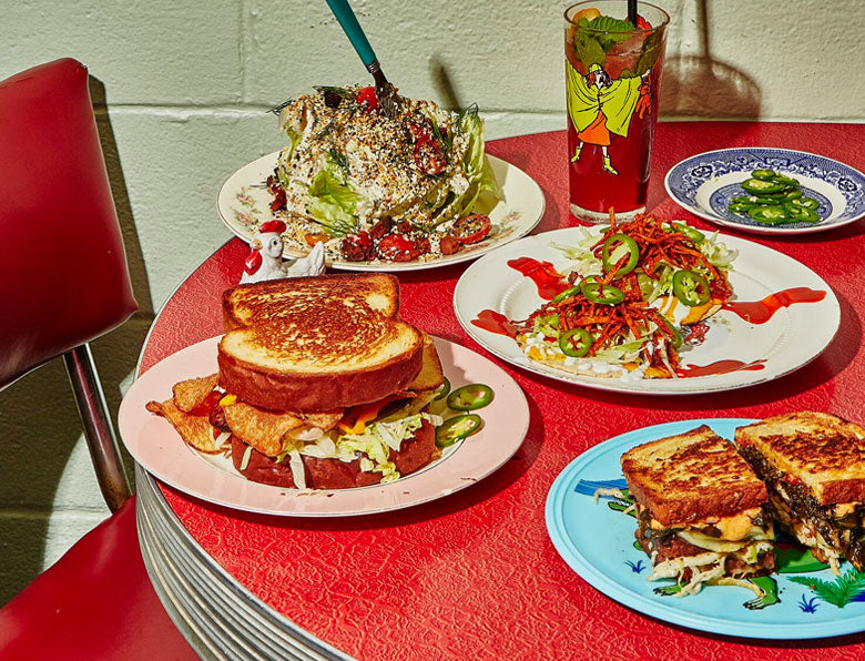 A diner-style table with a variety of sandwiches and salads, a drink with a straw, on red patterned tablecloth