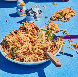 A bowl of shredded salad with utensils on a bright blue table, in sunny outdoor setting.