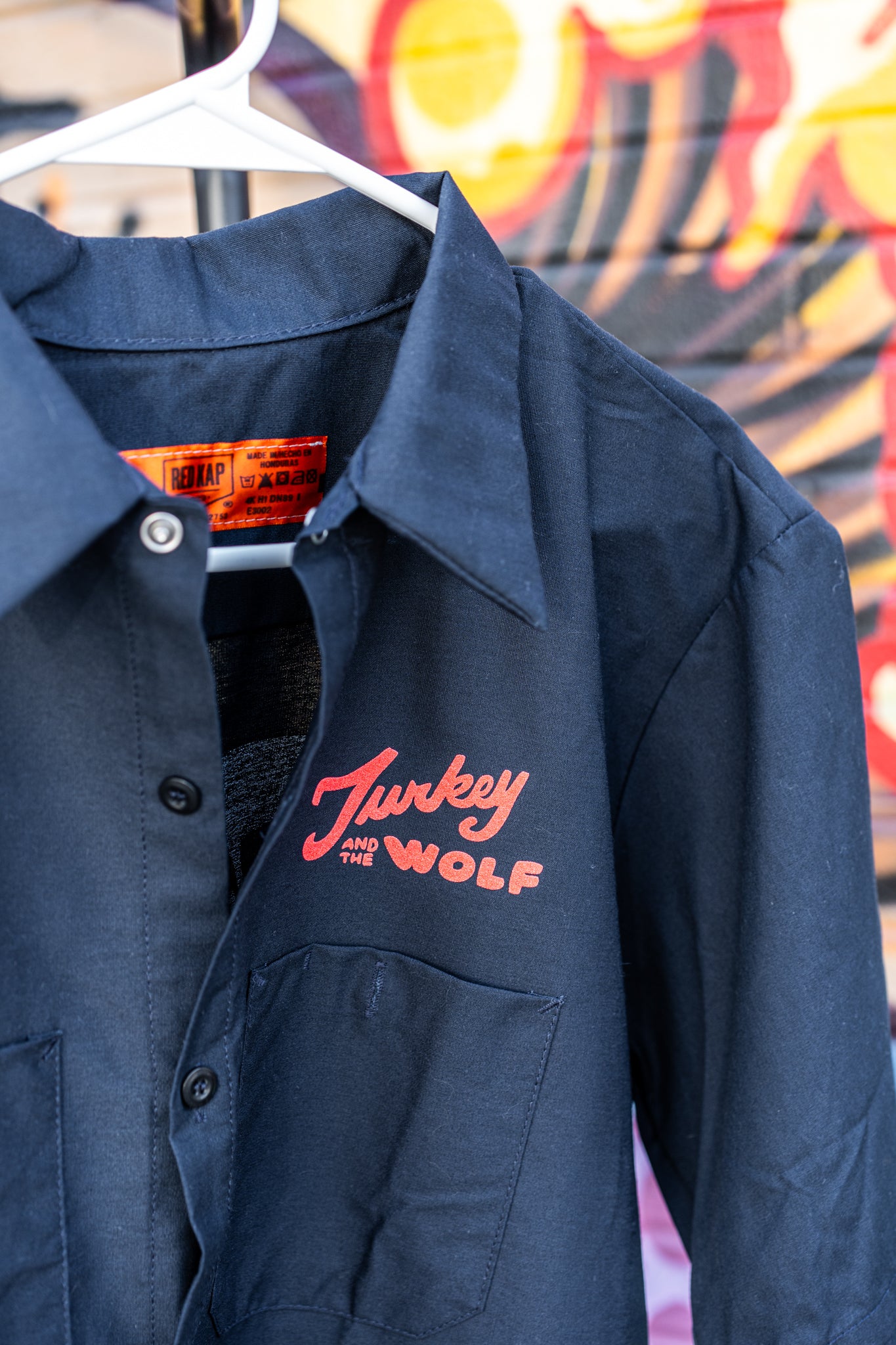 Navy blue shirt with red logo on a hanger against a colorful mural background