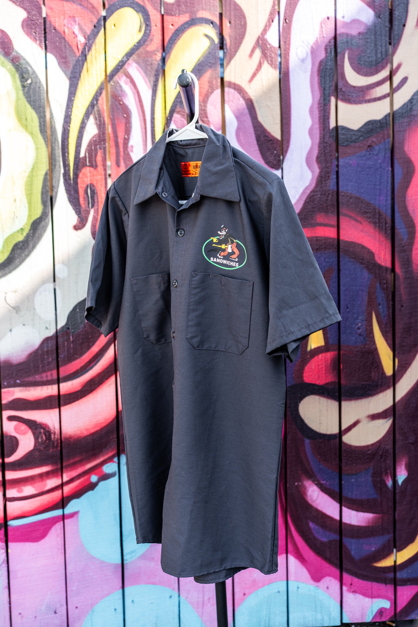 Black polo shirt with a logo on a hanger against a colorful mural background