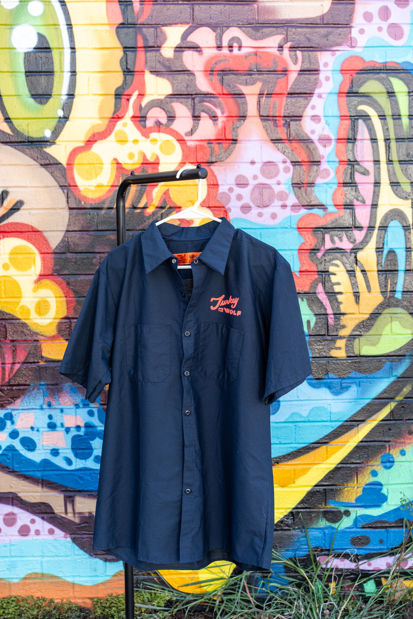 Navy blue shirt with a logo on a hanger against a colorful graffiti wall.